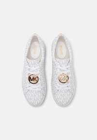 Zapatillas blancas con parte superior de tela que presenta un sutil patrón de logotipos de MK, cordones blancos y un emblema dorado de MK en la lengüeta.