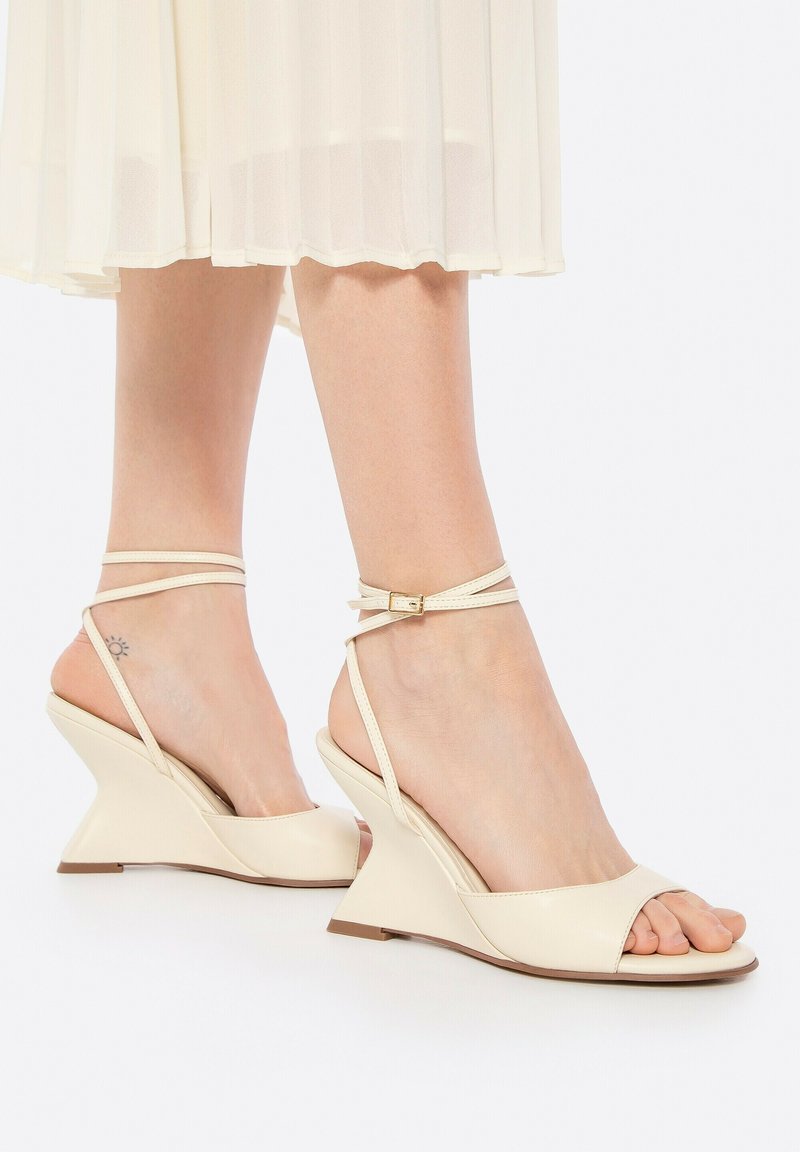 Sandales compensées beiges avec orteils ouverts et fines brides de cheville. Matériau lisse et brillant avec une forme moderne et angulaire. Détails de couture subtils.