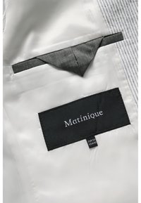 Svart tygmärke med vit text "Matinique", sydd på en slät vit innerfoder. Inkluderar en liten grå fickdetalj.