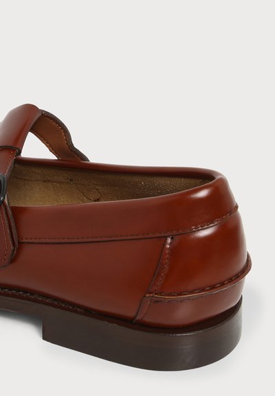 Mocassin en cuir marron avec un petit talon, des coutures visibles et une bride sur le dessus avec une boucle noire.