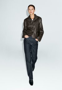 Donna che cammina in avanti indossando una giacca di pelle marrone scuro, jeans blu scuro e scarpe nere a punta con le mani nelle tasche.