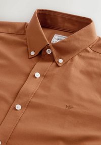 Pruun puuvillane nööpidega särk klassikalise kraega, valged nööbid ja väike broderie logo. Sile tekstuur, voolujoonis disain.