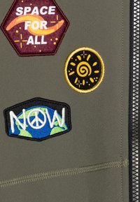 Tissu vert avec fermeture éclair présentant trois écussons brodés colorés : "ESPACE POUR TOUS", un soleil en spirale jaune et "MAINTENANT" avec un symbole de paix.