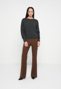 Dunkelgrauer Strickpullover mit weitem Ausschnitt, kombiniert mit braunen Schlaghosen. Das Modell trägt schwarze spitze High Heels. Einfarbiger Hintergrund.