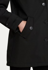 Manteau noir croisé à double boutonnage présentant un design ajusté avec de grands boutons, une texture lisse et des poches latérales. Style épuré et minimal.
