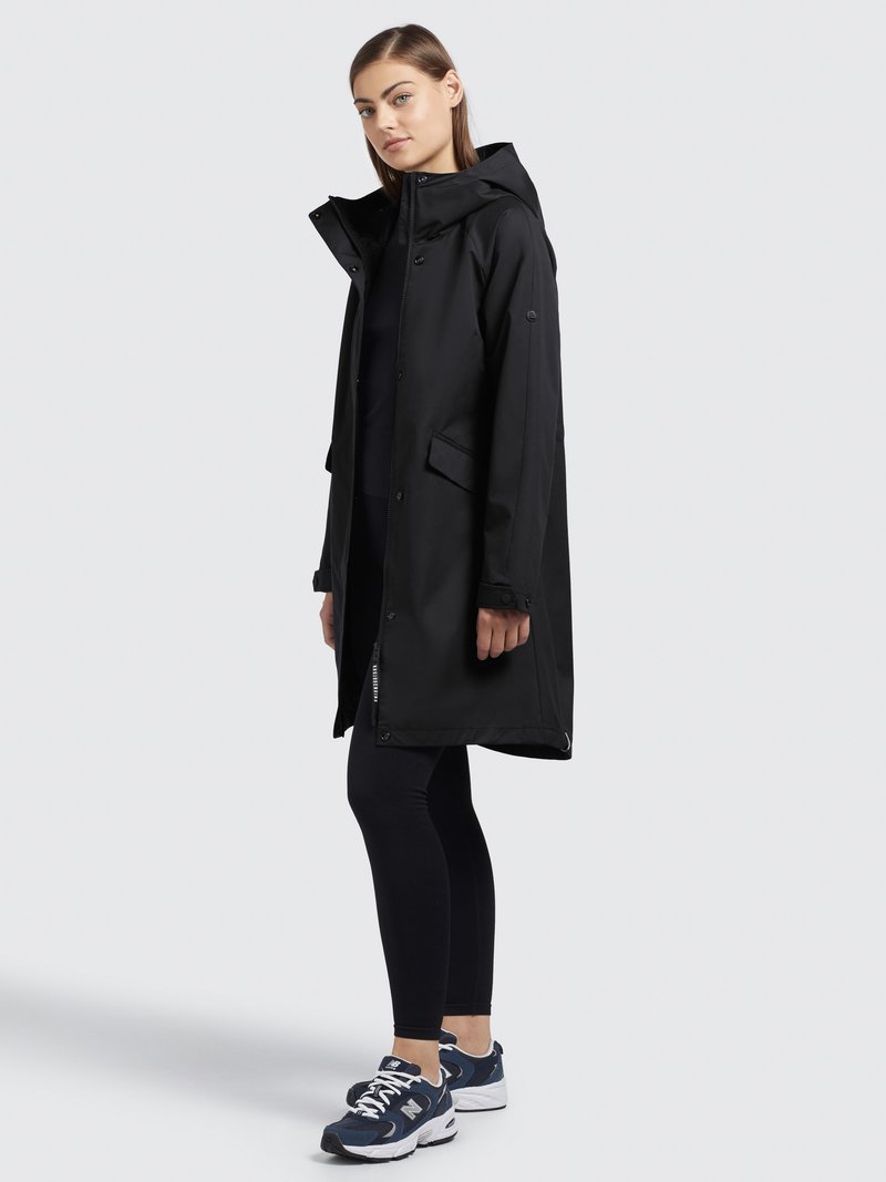 Manteau noir imperméable avec capuche, poches latérales et fermeture éclair. Associé à des leggings noirs et des baskets sportives. Texture lisse.