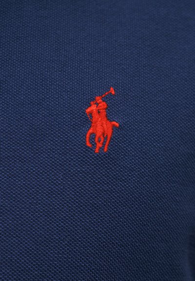 Giocatore di polo ricamato in rosso a cavallo che tiene un mazzuolo, cucito su tessuto blu scuro testurizzato.