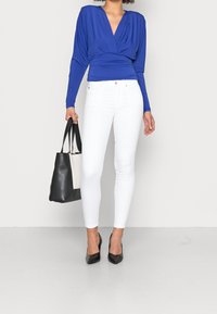 Blå långärmad wrap-topp med draperad design, ihopparad med vita skinny jeans. Svart tote-väska och spetsiga svarta klackar fulländar outfiten.