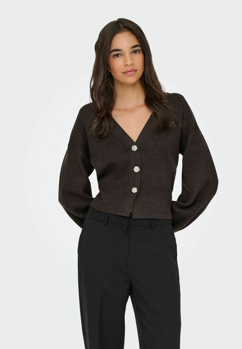 Brown cropped cardigan with a V-neck, long sleeves, and silver flower-shaped buttons. Worn over black trousers against a neutral background.