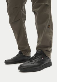 Čierne kožené tenisky s hladkým povrchom, okrúhlou špičkou, plochými šnúrkami a štruktúrovanou podrážkou. Kombinované s olivovými nohavicami, prezentujúce ležérny vzhľad.