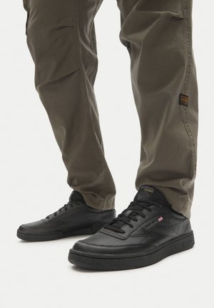 Čierne kožené tenisky s hladkým povrchom, okrúhlou špičkou, plochými šnúrkami a štruktúrovanou podrážkou. Kombinované s olivovými nohavicami, prezentujúce ležérny vzhľad.