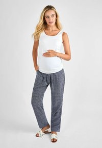 Top senza maniche bianca con scollo a barca abbinata a pantaloni blu larghi con un motivo a diamante. Il modello è scalzo e tiene delicatamente un pancione.
