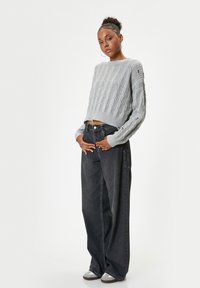 Grijze gebreide trui met ribbelstructuur en brede mouwen, gecombineerd met hoge, losse zwarte jeans en grijze sneakers.