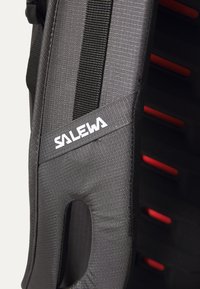Czarny materiałowy pasek plecaka z czerwonymi akcentami i teksturowaną powierzchnią. Posiada ergonomiczne wycięcie oraz detal z białym logo. Solidna konstrukcja zapewniająca trwałość.