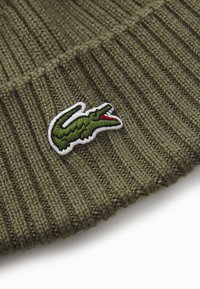 Bonnet en tricot vert olive avec texture côtelée et logo de crocodile brodé en contraste, en blanc et vert, avec des accents rouges.