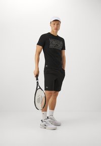 Musta t-paita, jossa on graafinen printti, mustat sortsit, valkoiset urheilukengät ja valkoinen lippalakki; pitää tennismailaa, jossa on verkkomuotoinen rakenne.