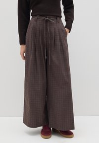 Pantalones de tiro ancho de cuadros marrones con pliegues y cintura con cordón, combinados con zapatillas de deporte burdeos, sobre un fondo blanco liso.