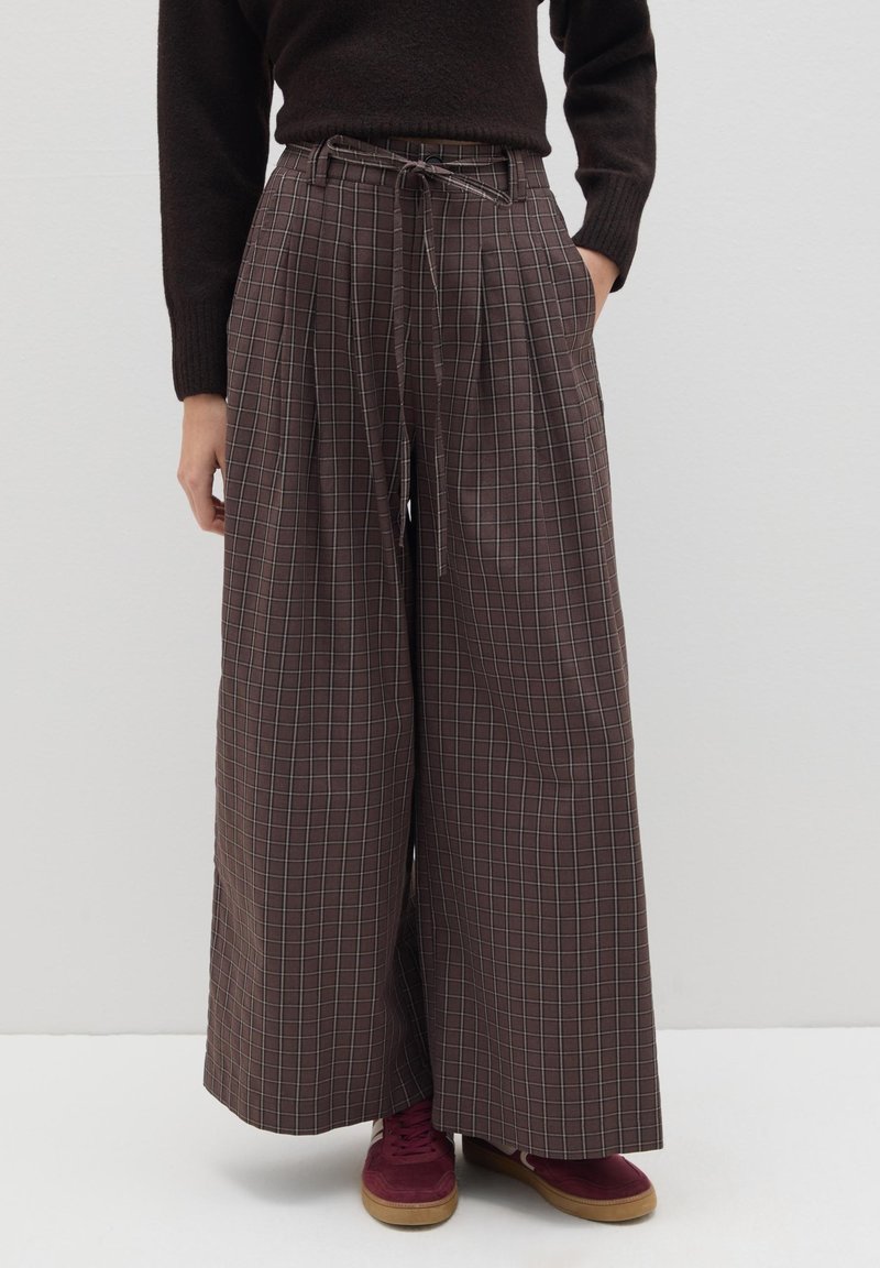 Pantalones de tiro ancho de cuadros marrones con pliegues y cintura con cordón, combinados con zapatillas de deporte burdeos, sobre un fondo blanco liso.