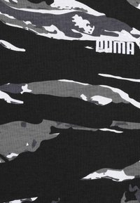 Textilie s camo vzorem, obsahující odstíny černé a šedé s bílými akcenty. Zahrnuje vyšívané logo Puma v bílé barvě. Texturovaná bavlněná směs.