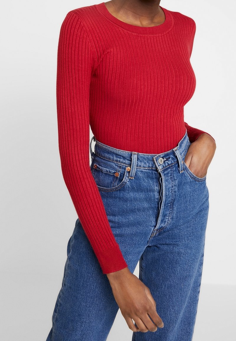 Femme portant un pull rouge côtelé ajusté rentré dans un jean bleu taille haute, main dans la poche, sur fond uni.