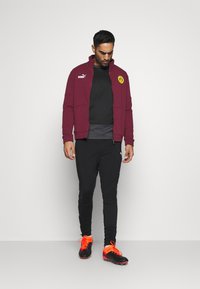 Burgundowa kurtka zapinana na zamek z logo Puma i żółtą łatą Borussia Dortmund, nałożona na czarną koszulę, zestawiona z czarnymi spodniami i piłkarskimi butami w kolorze czarnym/pomarańczowym.