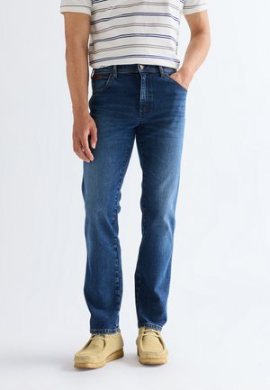 Mann trägt blaue Jeans, beigefarbene Schuhe und weiß gestreiftes kurzärmeliges Hemd, steht mit einer Hand in der Tasche vor schlichtem Hintergrund.