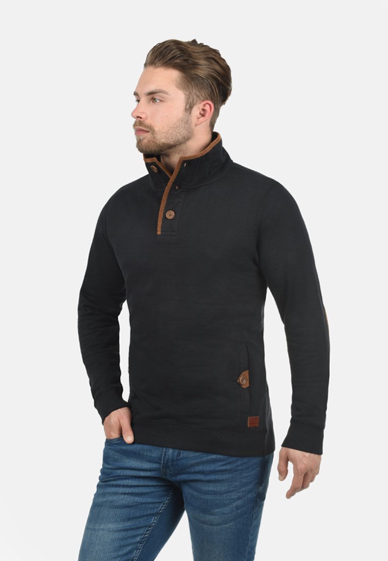 Schwarzer Pullover mit hohem Kragen, der braune Knopf-Details und eine Tasche mit einem Lederlogo aufweist. Hergestellt aus einem weichen, strukturierten Stoff.