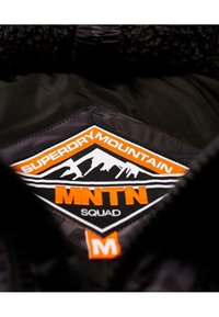 Svart innerfoder på en jacka med en broderad lapp. Lappen inkluderar texten "SUPERDRY MOUNTAIN," "MNTN," och "SQUAD" i orange och vitt.