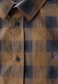 Hnědo-černá kostkovaná košile s knoflíkovým zapínáním, límečkem a texturovanou látkou. Na spodním pravém rohu se nachází černé logo.