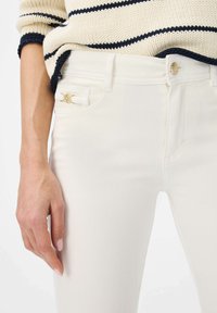 Witte denim broek heeft een gladde textuur met een aansluitend ontwerp. Een gouden accentdetail is zichtbaar op de zak, wat de algehele uitstraling verbetert.