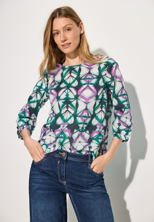 Camicia a maniche lunghe con un vivace motivo geometrico in verde, viola e nero, abbinata a jeans blu. Tessuto morbido e leggero.