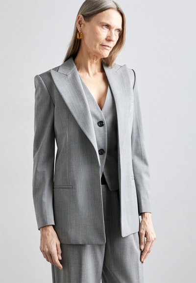 Costume gris à rayures avec un blazer structuré à décolleté en V profond et larges revers, associé à un gilet assorti et un pantalon ajusté.