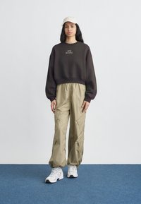 Γυναίκα που φοράει μαύρο φούτερ New Balance, μπεζ cargo παντελόνι, λευκά αθλητικά παπούτσια και εκρού καπέλο baseball, στέκεται σε μπλε χαλί μπροστά σε λευκό τοίχο.