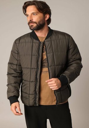 Veste bomber matelassée vert olive avec un col et des poignets côtelés noirs, fermeture éclair et poches latérales, superposée sur un t-shirt graphique marron.