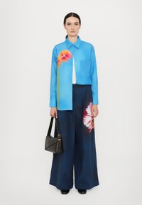 Blue long-sleeve shirt with floral print on left side, paired with wide-leg navy trousers, holding a black bag, on a plain background.