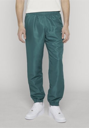 Hombre con pantalones jogger ajustables de cintura elástica en color verde azulado y zapatillas blancas, de pie contra un fondo liso y claro.