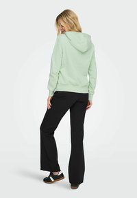 Felpa verde chiaro con cappuccio e cordino, abbinata a pantaloni neri a campana e scarpe nere con dettagli bianchi. Il tessuto è morbido e casual.