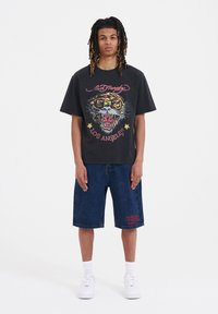 Black cotton t-shirt featuring a colorful tiger graphic and text on the front, paired with dark denim shorts and white sneakers.
