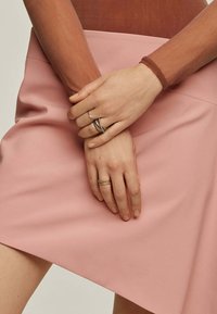 Falda rosa con una textura suave y un dobladillo en ángulo. Manos adornadas con múltiples anillos, con tonos de plata y oro. Blusa larga de manga larga de tono marrón transparente.