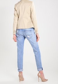Blazer in pelle beige con design aderente, abbinato a jeans strappati azzurri e décolleté nudi, con maniche arrotolate e finitura usurata.