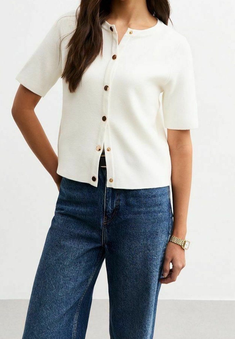 Femme portant un haut blanc à manches courtes boutonné et un jean bleu, debout avec une main dans la poche, montre dorée au poignet.