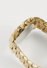 Montre dorée avec un boîtier rond lisse, des boutons-poussoirs texturés et un bracelet à maillons comportant des maillons rectangulaires.