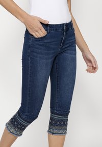 Un jean capri bleu foncé au design ajusté, avec des broderies décoratives et des clous au niveau de l'ourlet et des poches latérales.