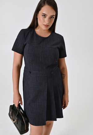 Dunkelblaues Jeanskleid mit kurzen Ärmeln, taillierter Oberpartie, plissiertem Rock, rundem Ausschnitt und seitlichen Knöpfen. Das Model hält eine schwarze Handtasche.