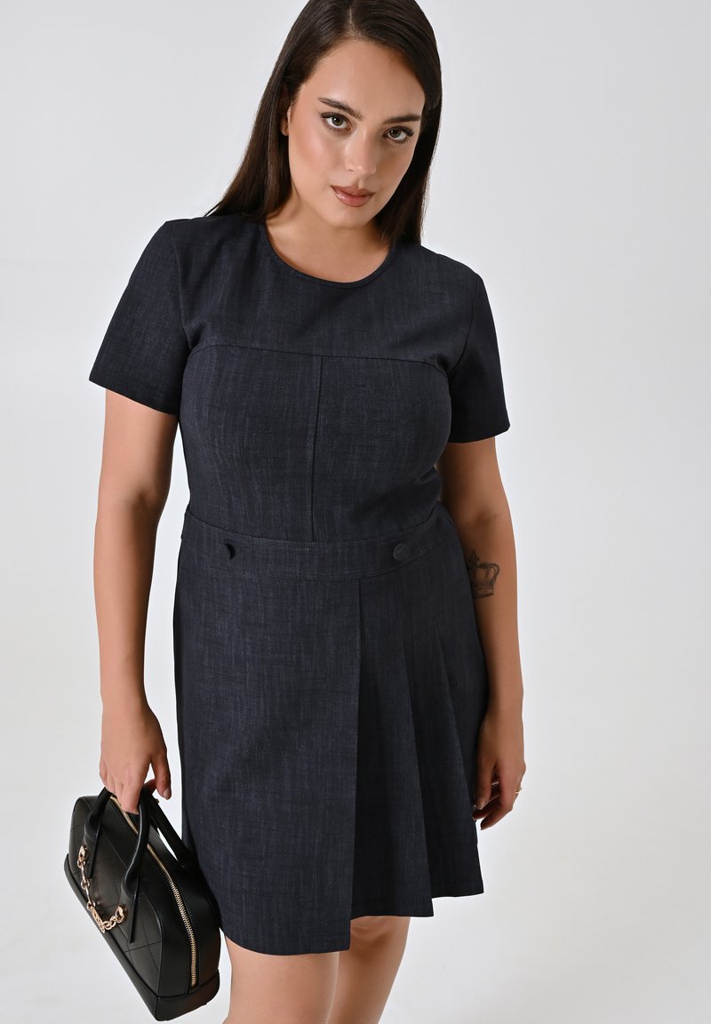 Robe en denim foncé à manches courtes avec un corsage ajusté, une jupe plissée, un col rond et des boutons sur le côté. Le mannequin tient un sac à main noir.