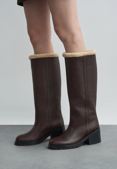 Brown leather knee-high boots with beige fleece lining, chunky black heel, and rubber sole; smooth texture with vertical seams.