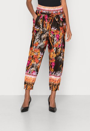 Femme portant un pantalon à motifs floraux noirs, orange, rose et blancs avec taille et poignets élastiques, associé à des talons hauts noirs.