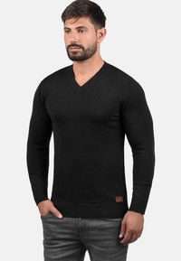 Maglione nero a maglia con scollatura a V e maniche lunghe, con polsini e orlo a coste, caratterizzato da una piccola etichetta in pelle marrone sul bordo inferiore.