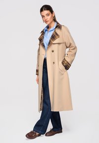 Femme portant un long manteau beige avec col et poignets à carreaux, une chemise bleu clair, un jean foncé et des chaussures marron, debout avec une main dans la poche.