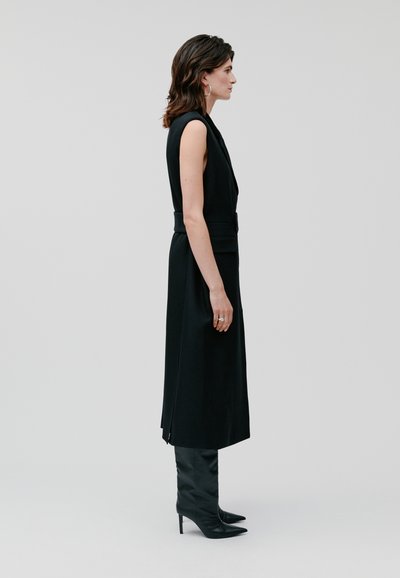 Zwarte mouwloze jurk met een getailleerde taille, zijsplitten en een gladde textuur. Gemaakt in combinatie met zwart leren laarzen tot de knie met hak.